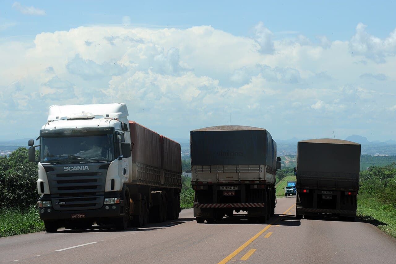 Três caminhões graneleiros com carga coberta por lona trafegam em uma rodovia de pista simples, sob céu com nuvens