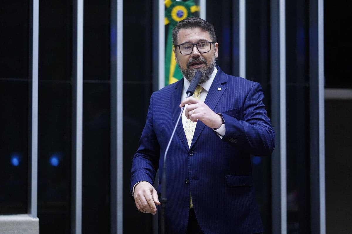 Deputado de terno azul e óculos fala ao microfone no plenário da Câmara