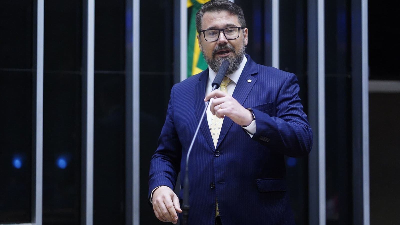 Deputado de terno azul e óculos fala ao microfone no plenário da Câmara