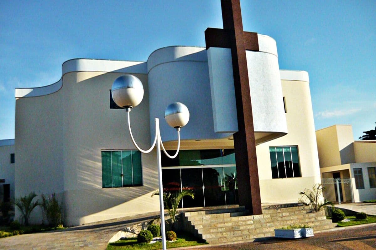 Fachada branca e curva de igreja moderna com grande cruz marrom e poste de luz com duas esferas à frente