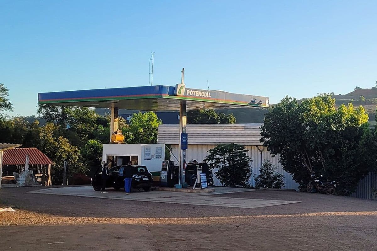 Posto de combustível pequeno em área rural, com cobertura azul, um carro abastecendo, galpão ao fundo e morros com vegetação