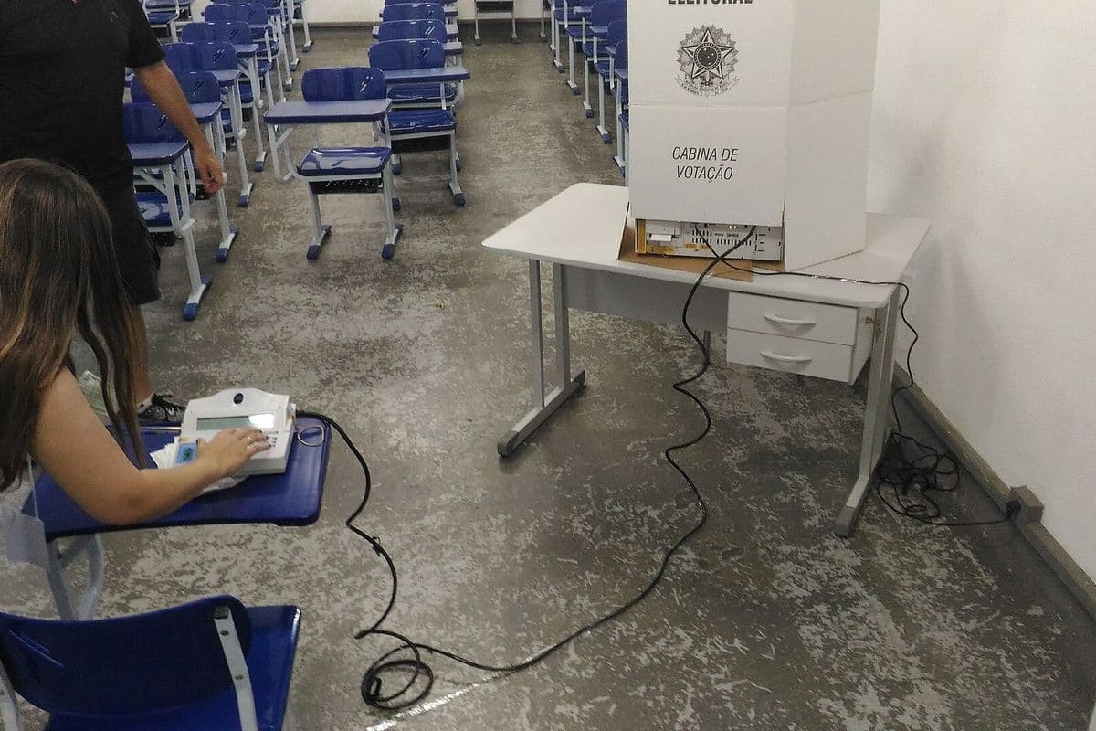 Mesária digita no terminal do mesário sobre uma carteira escolar, com a cabine de votação branca e a urna eletrônica sobre uma mesa ao fundo