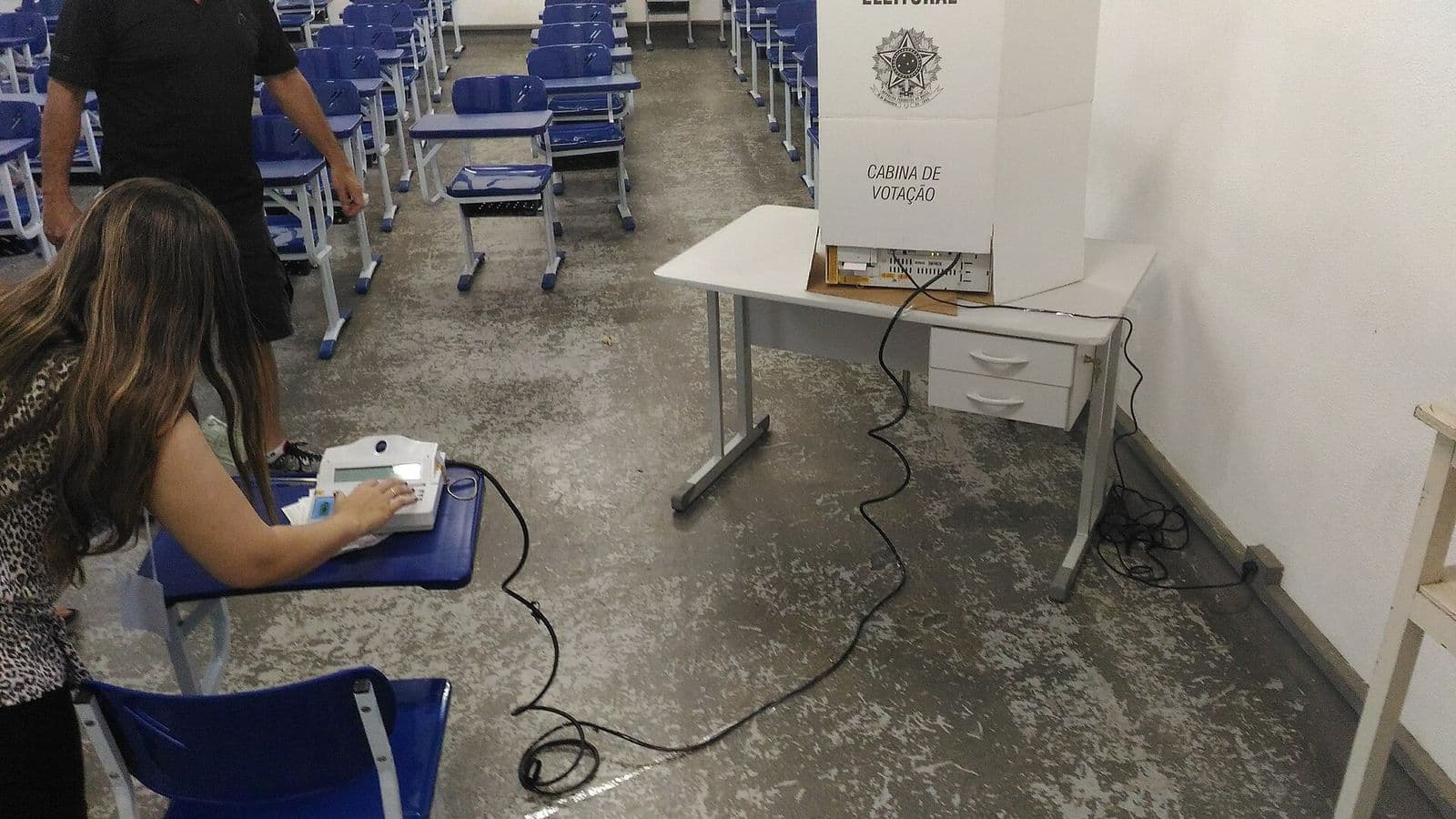 Mesária digita no terminal do mesário sobre uma carteira escolar, com a cabine de votação branca e a urna eletrônica sobre uma mesa ao fundo