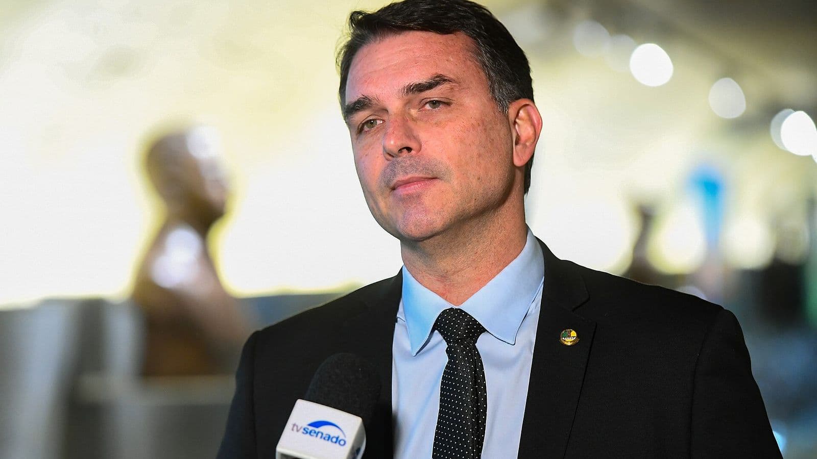 Flávio Bolsonaro, de terno escuro e gravata, fala diante de um microfone da TV Senado