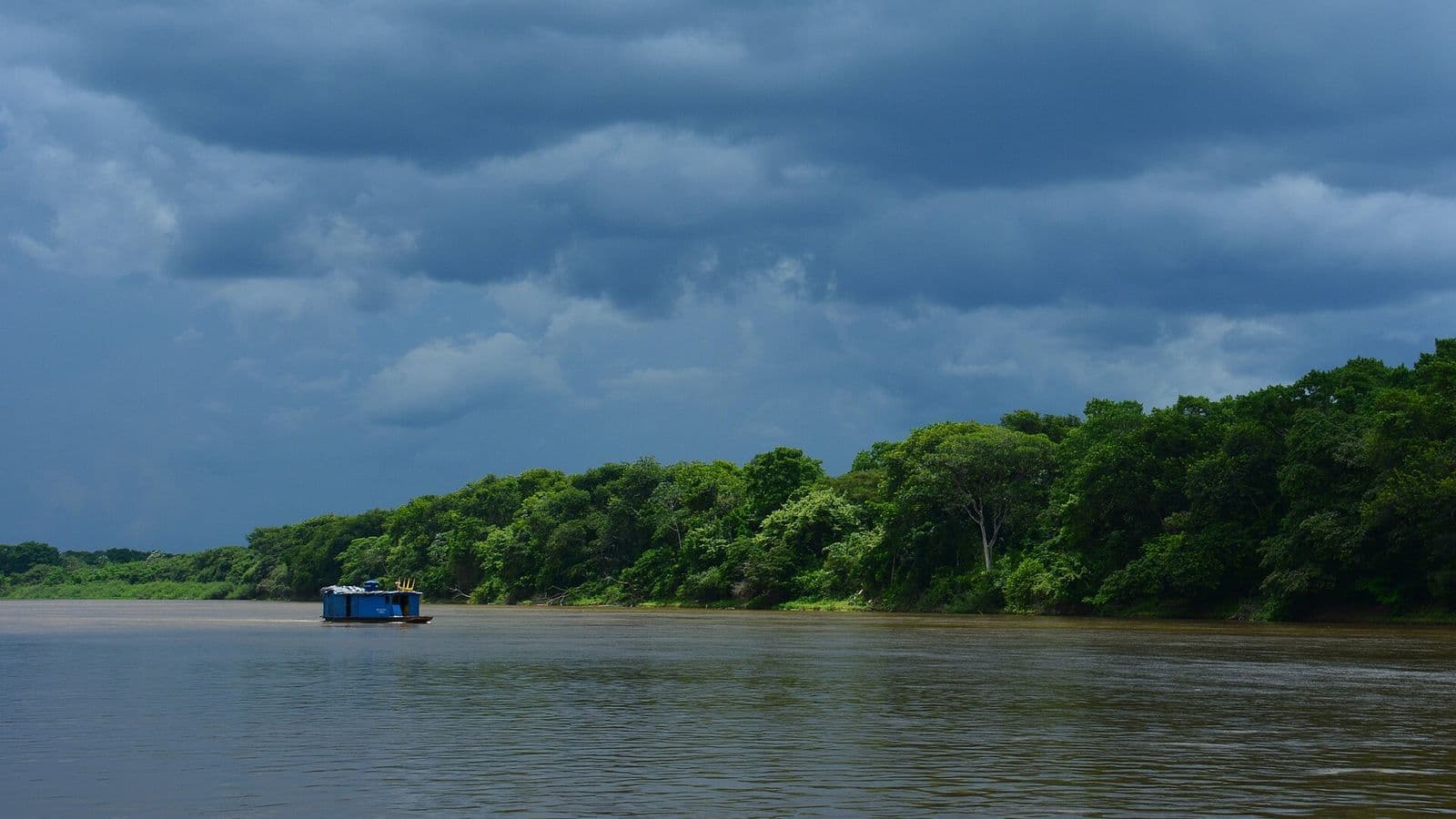 Nuvens escuras de tempestade cobrem o céu sobre um rio largo de água barrenta, com mata densa na margem e um pequeno barco azul