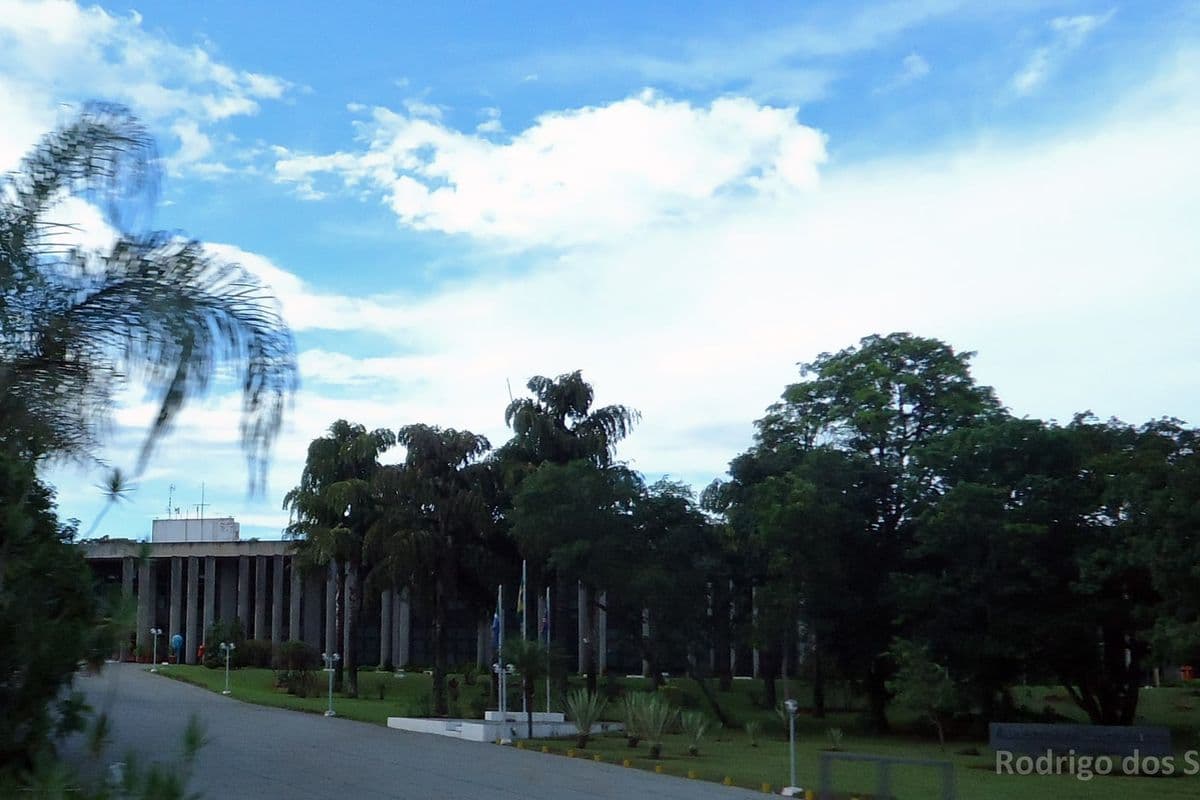 Prédio da Assembleia Legislativa cercado de árvores e palmeiras, sob céu azul com nuvens