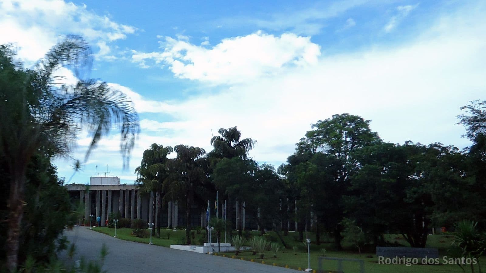 Prédio da Assembleia Legislativa cercado de árvores e palmeiras, sob céu azul com nuvens