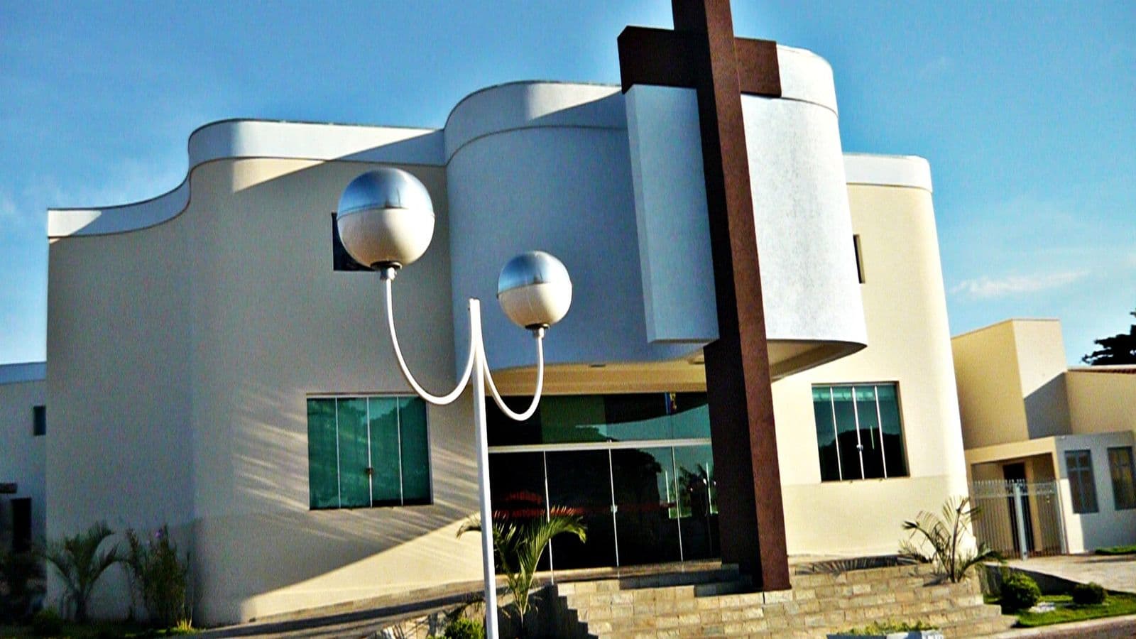 Fachada branca e curva de igreja moderna com grande cruz marrom e poste de luz com duas esferas à frente